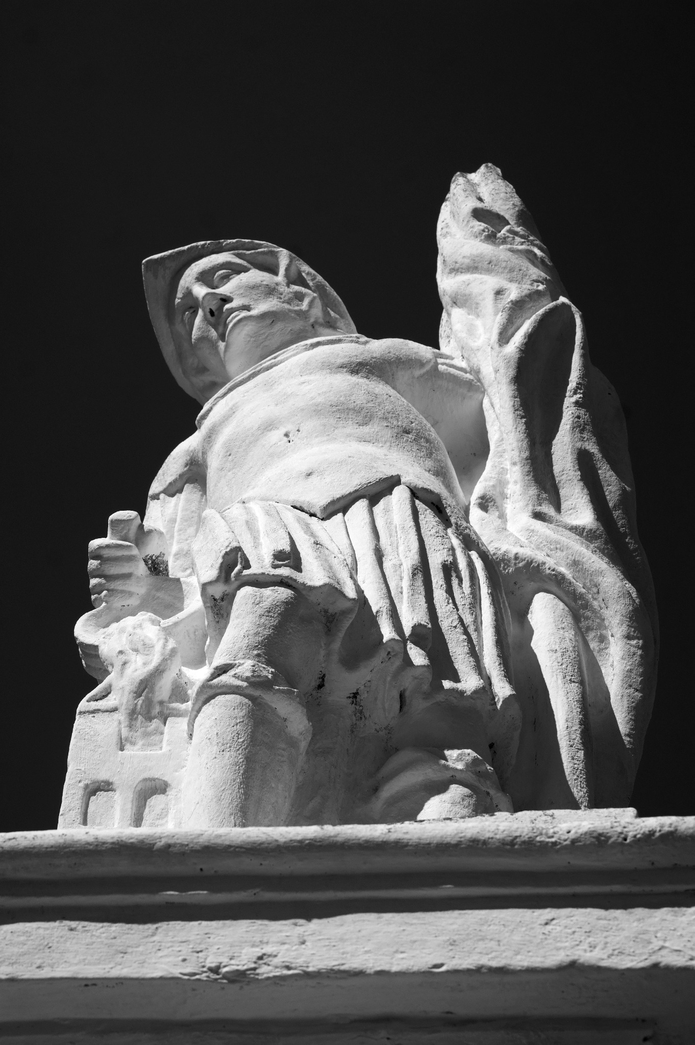 Czarno-białe zdjęcie z dołu przedstawiające kamienną rzeźbę postaci w szatach, z twarzą skierowaną ku górze i trzymającą w dłoniach coś w rodzaju zwoju lub księgi, na tle ciemnego nieba.