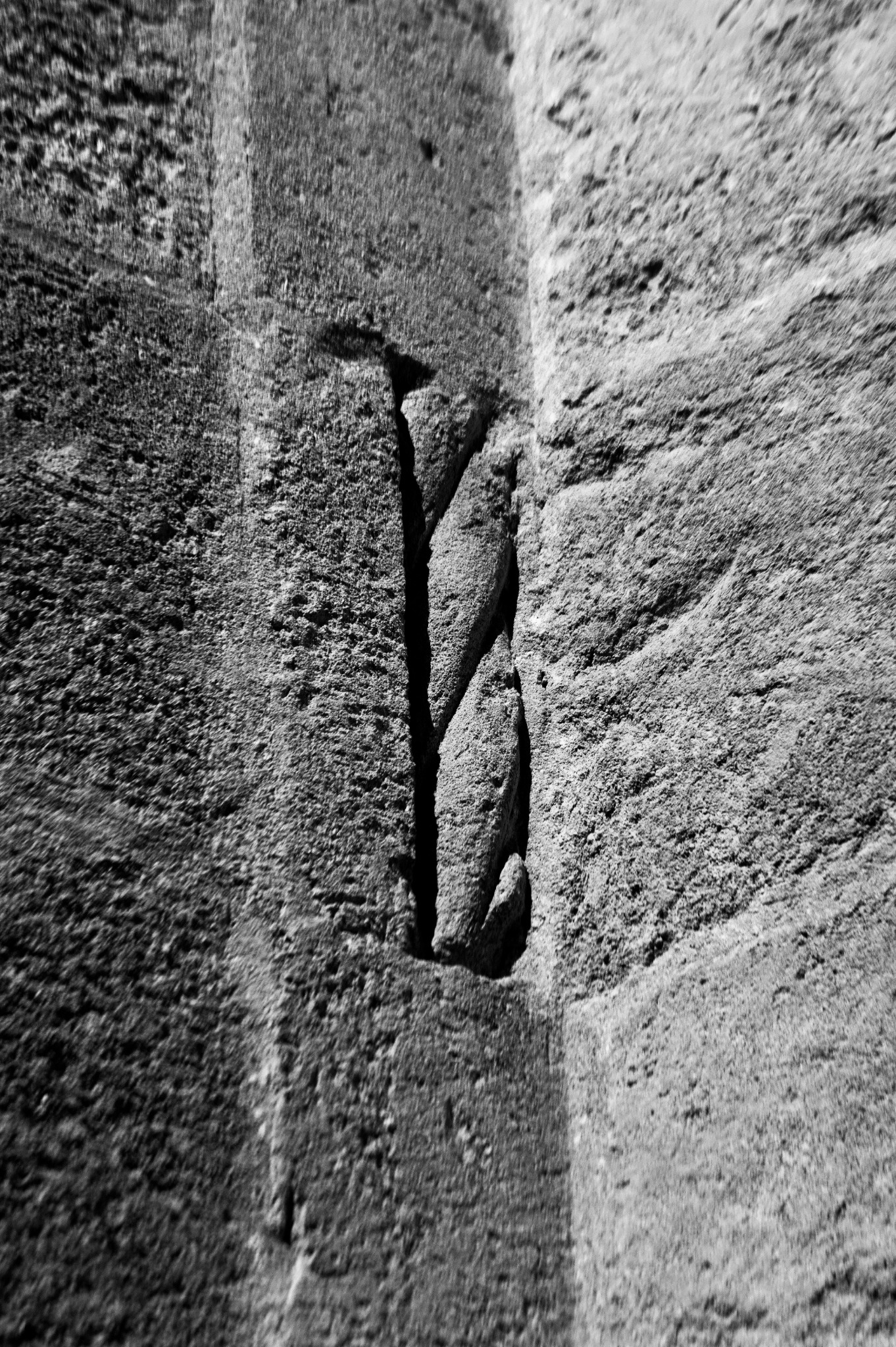 Czarno-białe zbliżenie fragmentu kamiennej ściany z wyraźnym pionowym pęknięciem, ukazującym teksturę i nierówności materiału.