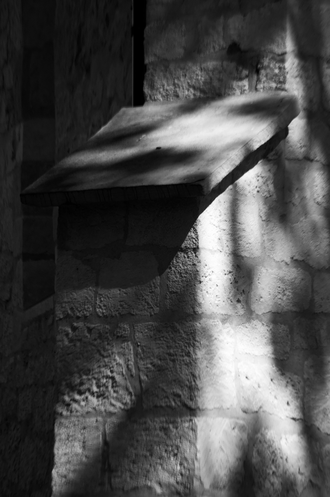 Czarno-białe zdjęcie fragmentu kamiennej ściany z wystającym kamiennym elementem, oświetlonej promieniami światła wpadającymi z góry.