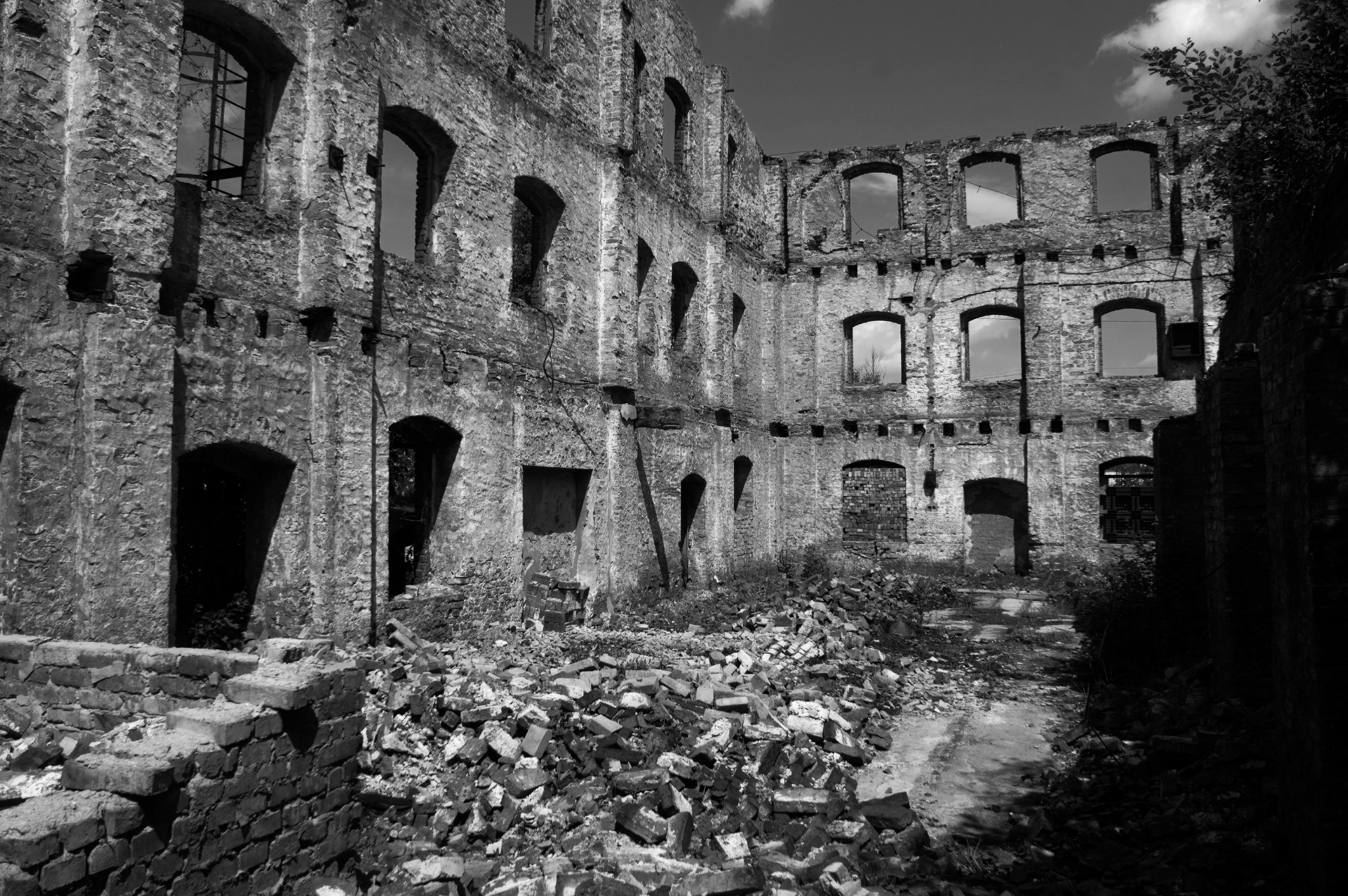 Czarno-białe zdjęcie ruin ceglanego budynku z licznymi oknami bez szyb i stertą gruzu na pierwszym planie, z widocznymi śladami rozbiórki.
