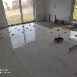 Białe płytki podłogowe z marmurkowym wzorem, ułożone w jasnym pomieszczeniu z dużymi oknami, podczas procesu poziomowania z użyciem czerwonych klinów i systemu poziomującego. W tle widoczne...