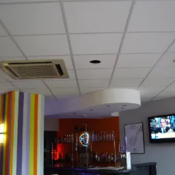 Kasetonowy sufit z kratką, oświetleniem punktowym i nawiewem klimatyzacji w lokalu z barem i telewizorem.