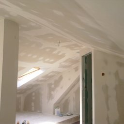 FRB IGMAR Mariusz Igielski - Poddasze w trakcie wykończenia: ściany i sufit pokryte szpachlą na płytach gipsowo-kartonowych, widoczne otwory na gniazdka elektryczne, okno dachowe i materiały budowlane.