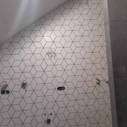 FRB IGMAR Mariusz Igielski - Ściana łazienki wyłożona białymi, sześciennymi płytkami z czarnymi krawędziami, widoczne niedokończone instalacje wodno-kanalizacyjne i elektryczne.