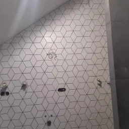 Ściana łazienki wyłożona białymi, sześciennymi płytkami z czarnymi krawędziami, widoczne niedokończone instalacje wodno-kanalizacyjne i elektryczne.