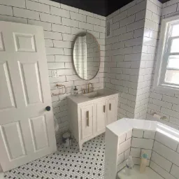 Wykończona łazienka z białymi, prostokątnymi płytkami na ścianach, czarnym sufitem, mozaikową podłogą w czarno-białe heksagony, owalnym lustrem w złotej ramie i białą szafką pod umywalką z złotymi...