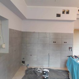 FRB IGMAR Mariusz Igielski - Wnętrze kuchni w trakcie wykończenia: szare płytki ścienne, białe ściany, przygotowane do malowania, widoczne puszki z farbą i zabezpieczone blaty.