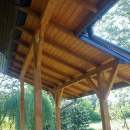 Drewniana konstrukcja zadaszenia tarasu z widocznym systemem rynnowym i elementami mocującymi, na tle zieleni drzew, z logo firmy FRB IGMAR w prawym górnym rogu.