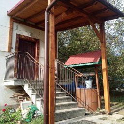 FRB IGMAR Mariusz Igielski - Zadaszone wejście do domu z drewnianą konstrukcją, metalową balustradą i studnią z wiadrem w tle.