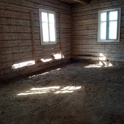 FRB IGMAR Mariusz Igielski - Wnętrze starego domu z drewnianymi ścianami, ceglanym fundamentem i ziemistą podłogą, oświetlone promieniami słońca wpadającymi przez okna.