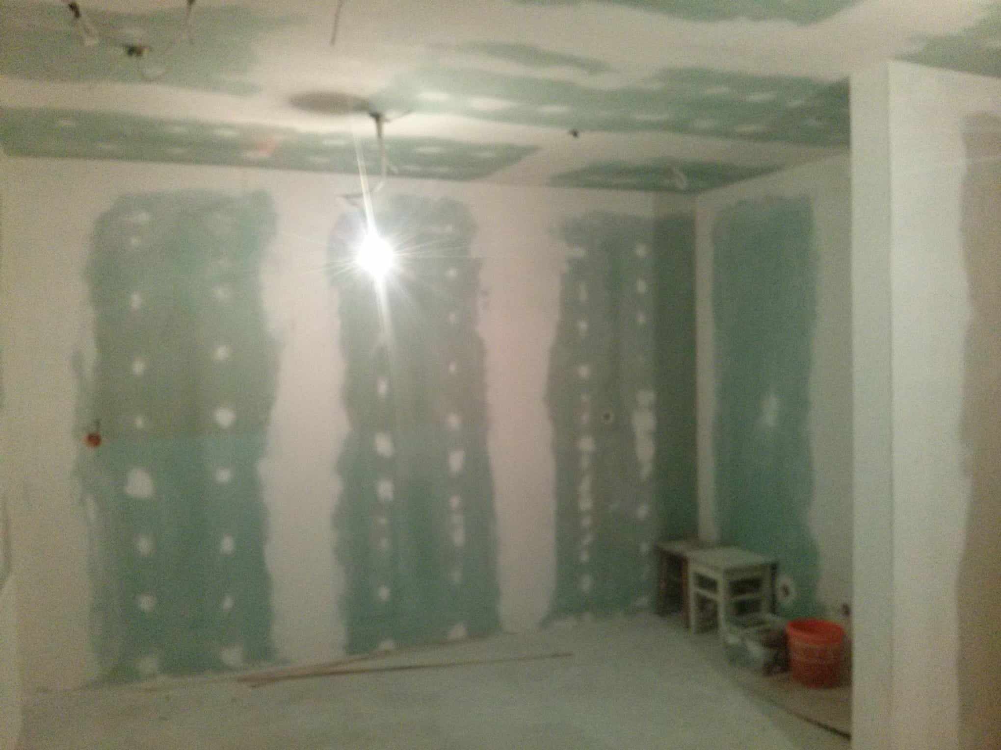 Wnętrze w trakcie prac wykończeniowych: widoczne płyty kartonowo-gipsowe na ścianach i suficie, szpachlowane łączenia, prowizoryczne oświetlenie i narzędzia budowlane w tle.