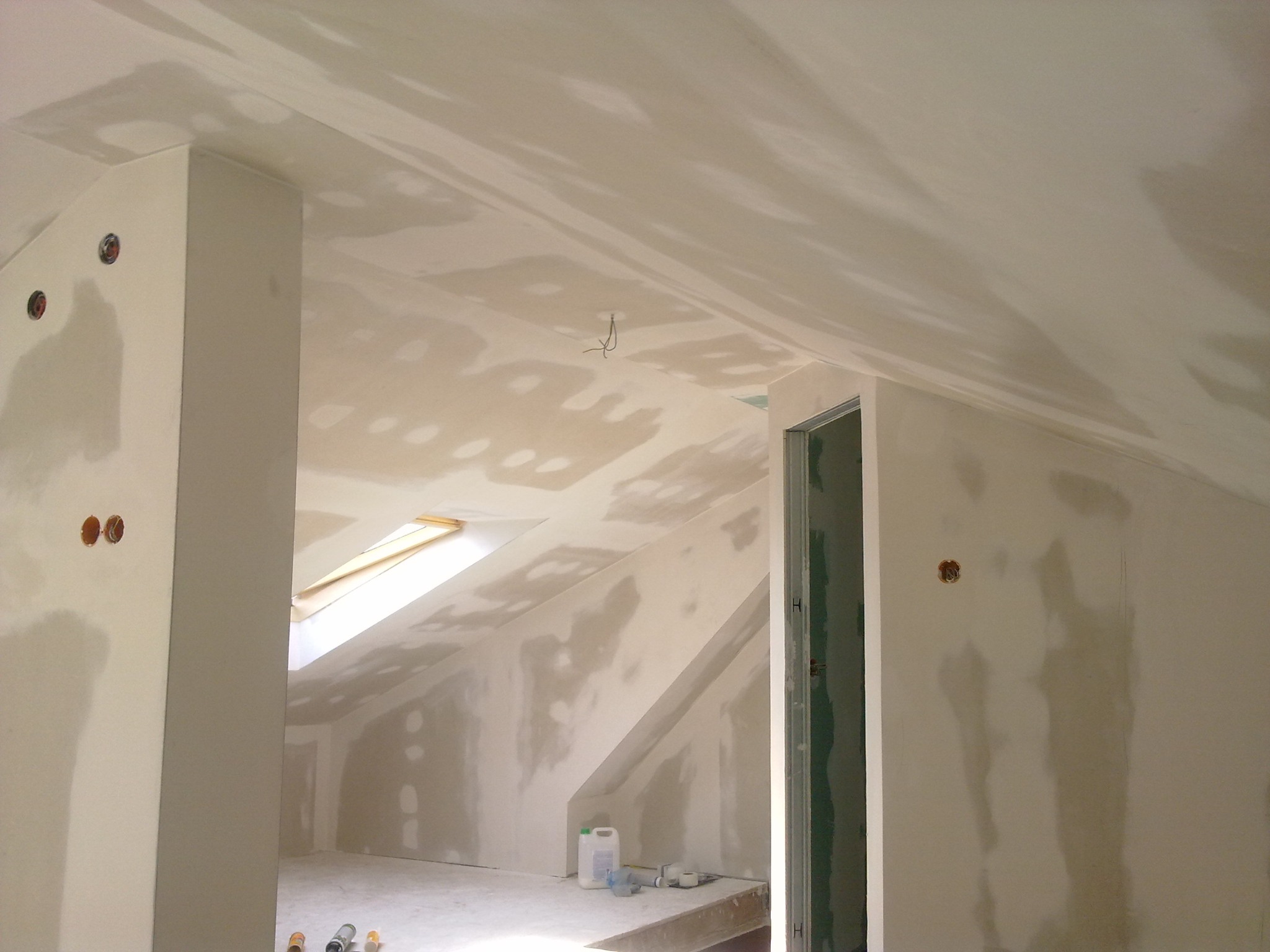 Poddasze w trakcie wykończenia: ściany i sufit pokryte szpachlą na płytach gipsowo-kartonowych, widoczne otwory na gniazdka elektryczne, okno dachowe i materiały budowlane.