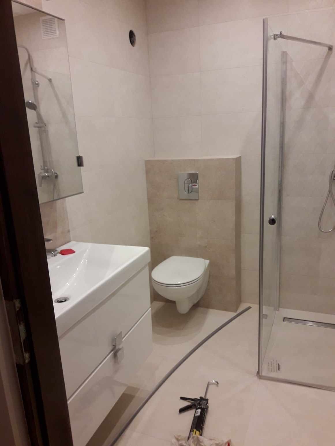 Nowoczesna łazienka z podwieszaną toaletą, białą umywalką z szafką i kabiną prysznicową, widoczny aplikator do silikonu na podłodze, beżowe płytki na ścianach.