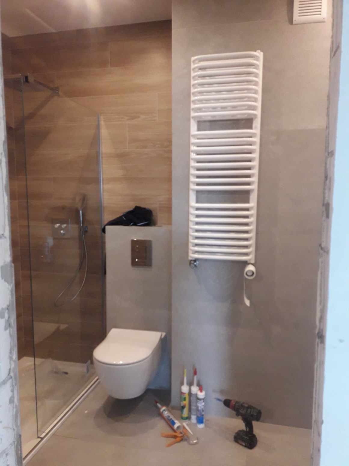 Nowoczesna łazienka w trakcie wykańczania: widoczna kabina prysznicowa z drewnopodobnymi płytkami, podwieszana toaleta z czarnym obuwiem na spłuczce, grzejnik drabinkowy na szarej ścianie...