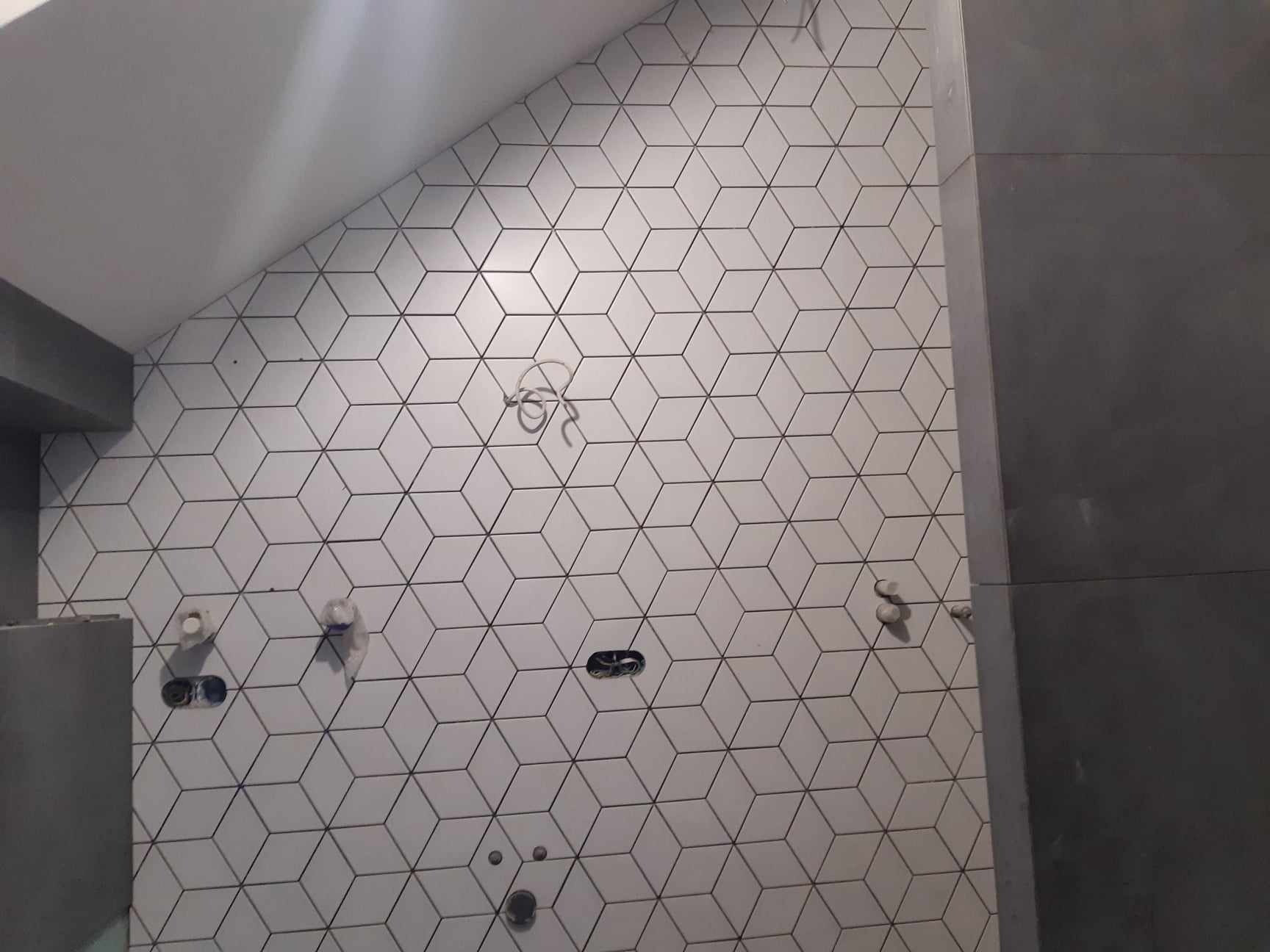 Ściana łazienki wyłożona białymi, sześciennymi płytkami z czarnymi krawędziami, widoczne niedokończone instalacje wodno-kanalizacyjne i elektryczne.