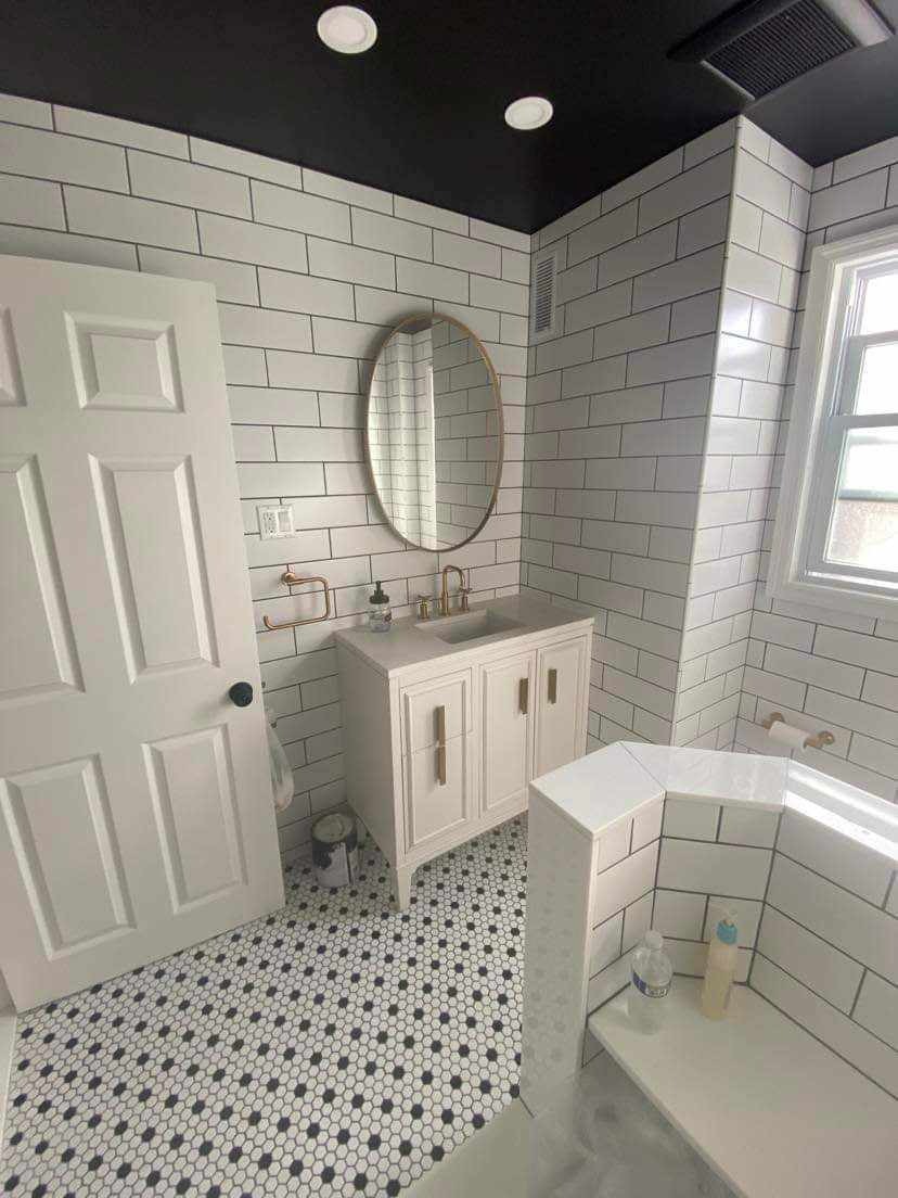 Wykończona łazienka z białymi, prostokątnymi płytkami na ścianach, czarnym sufitem, mozaikową podłogą w czarno-białe heksagony, owalnym lustrem w złotej ramie i białą szafką pod umywalką z złotymi...