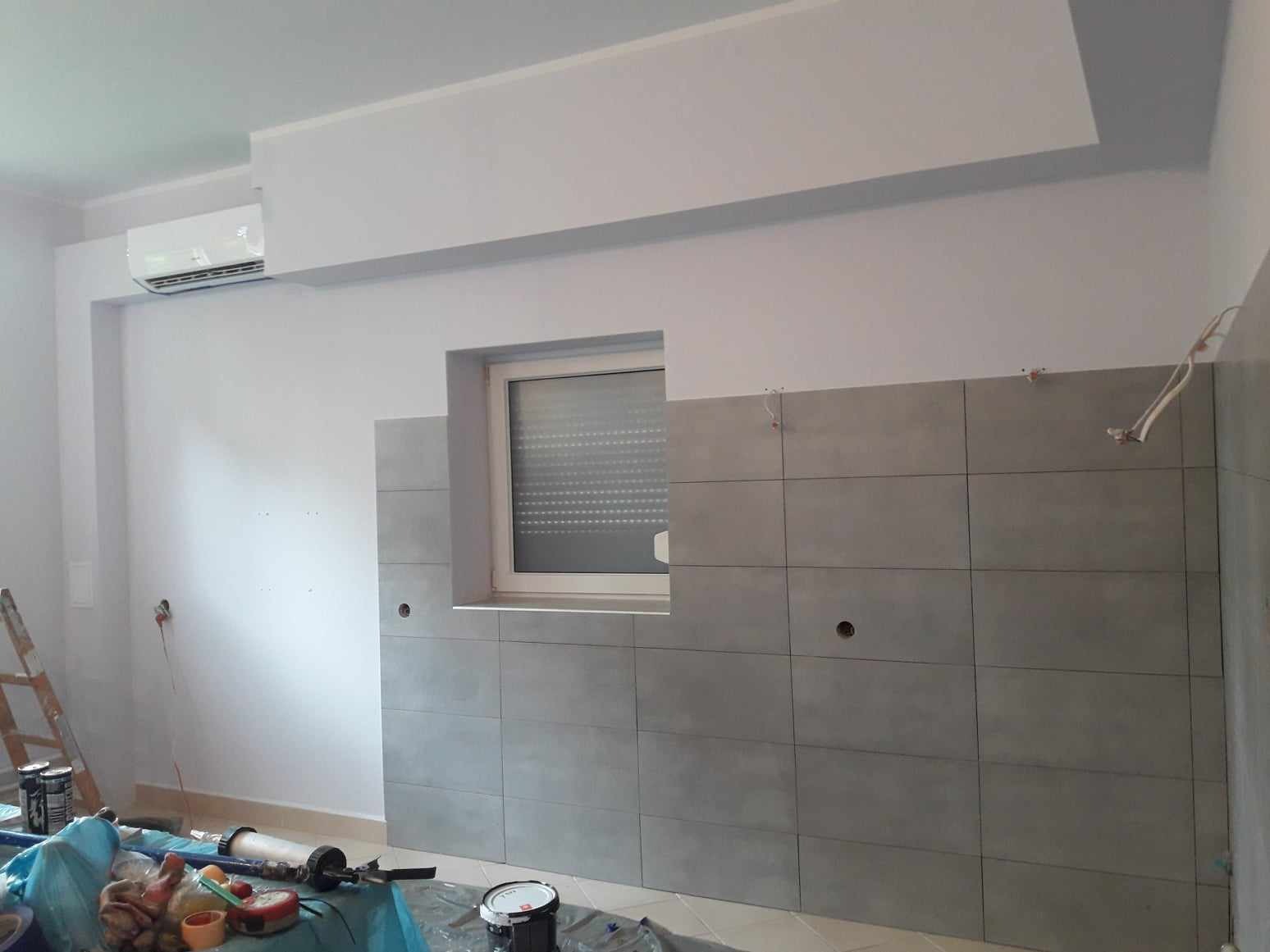 Montaż szarych, prostokątnych płytek ściennych w pomieszczeniu z oknem; widoczne przewody elektryczne, narzędzia i drabina.  Płytki ułożone do połowy ściany, z wywierconymi otworami.