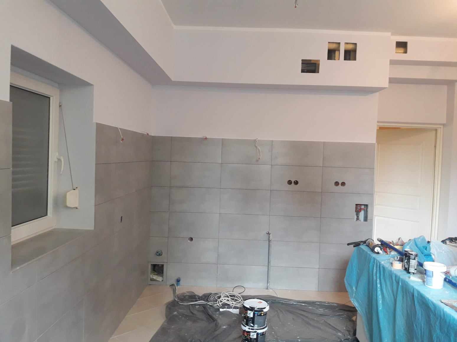 Wnętrze kuchni w trakcie wykończenia: szare płytki ścienne, białe ściany, przygotowane do malowania, widoczne puszki z farbą i zabezpieczone blaty.