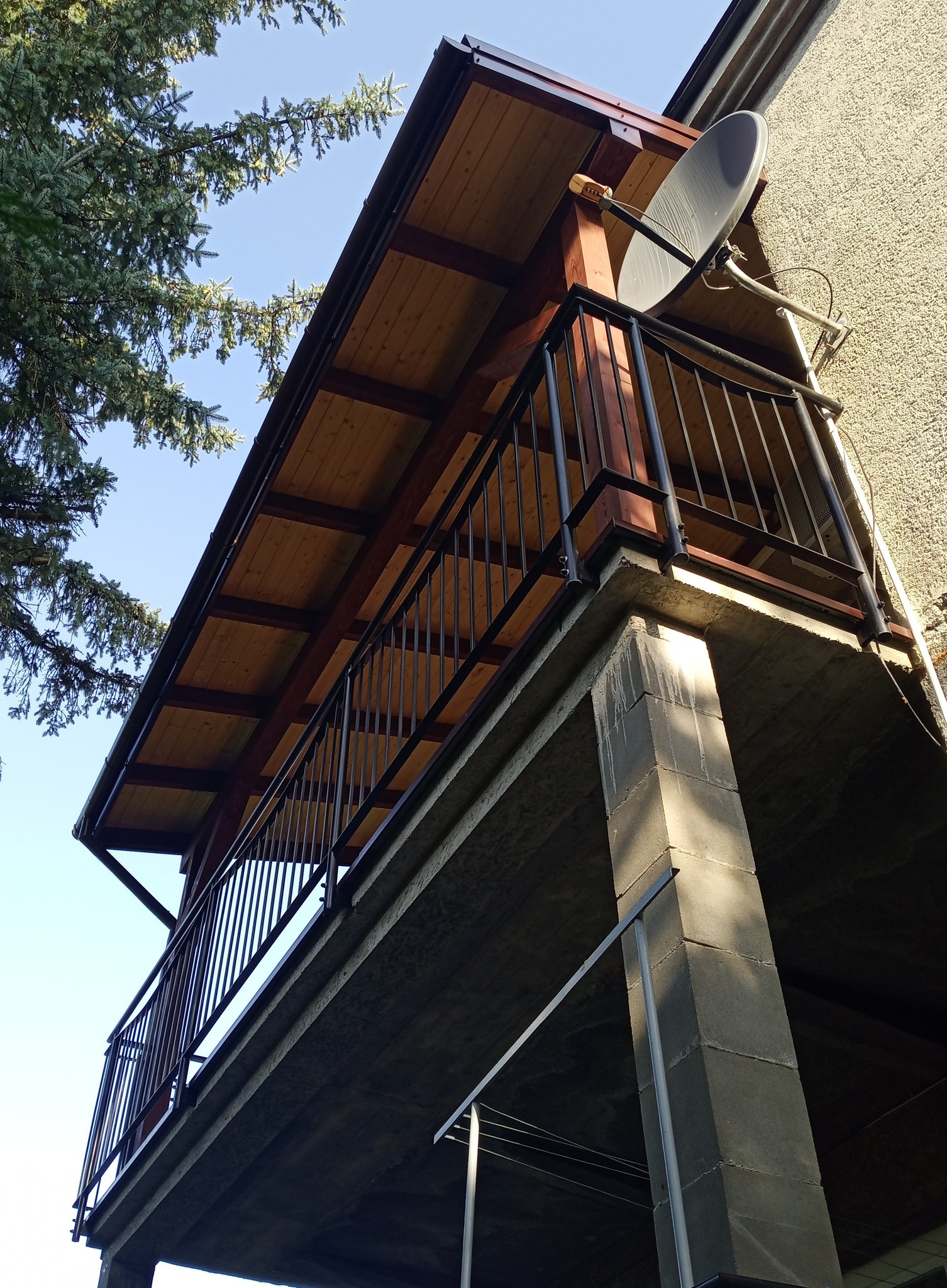 Zadaszony balkon z drewnianym sufitem i metalową balustradą, widok z dołu, z anteną satelitarną na ścianie budynku.