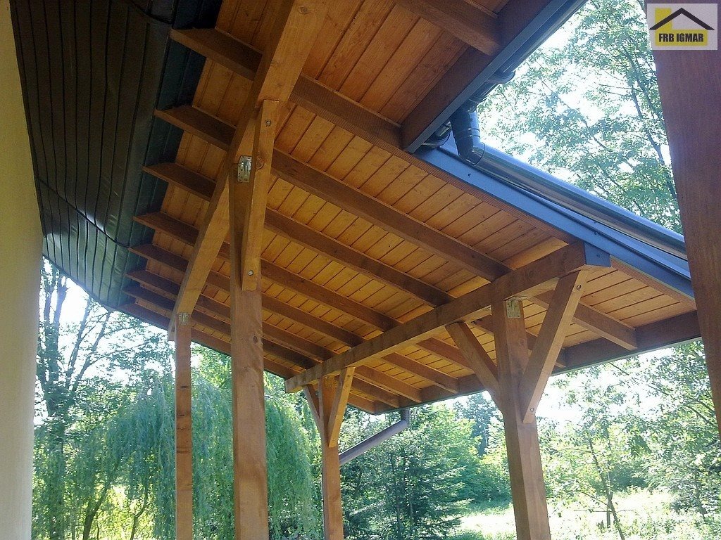 Drewniana konstrukcja zadaszenia tarasu z widocznym systemem rynnowym i elementami mocującymi, na tle zieleni drzew, z logo firmy FRB IGMAR w prawym górnym rogu.
