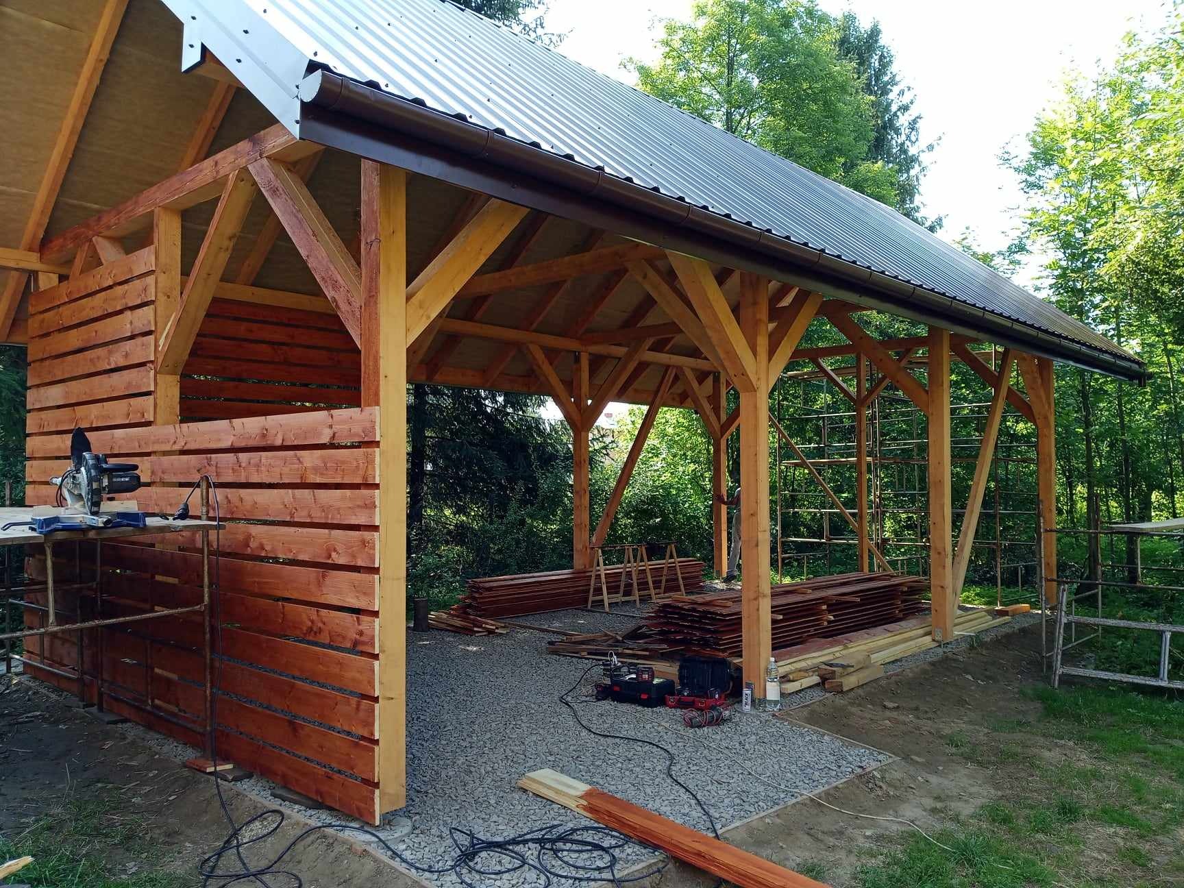 Drewniana konstrukcja z częściowo ukończonymi ścianami z desek, metalowym dachem i brązowymi rynnami, otoczona zielenią, z narzędziami i materiałami budowlanymi na żwirowym podłożu.