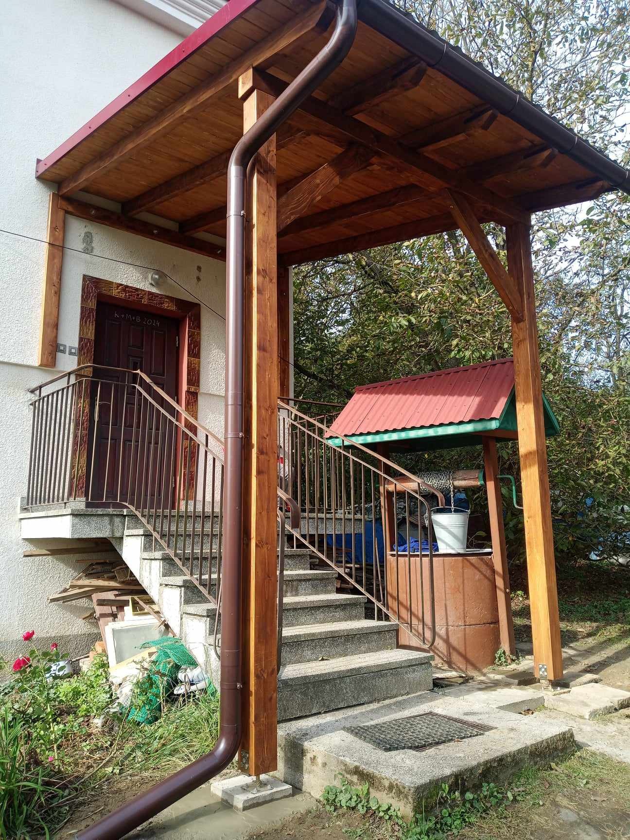 Zadaszone wejście do domu z drewnianą konstrukcją, metalową balustradą i studnią z wiadrem w tle.