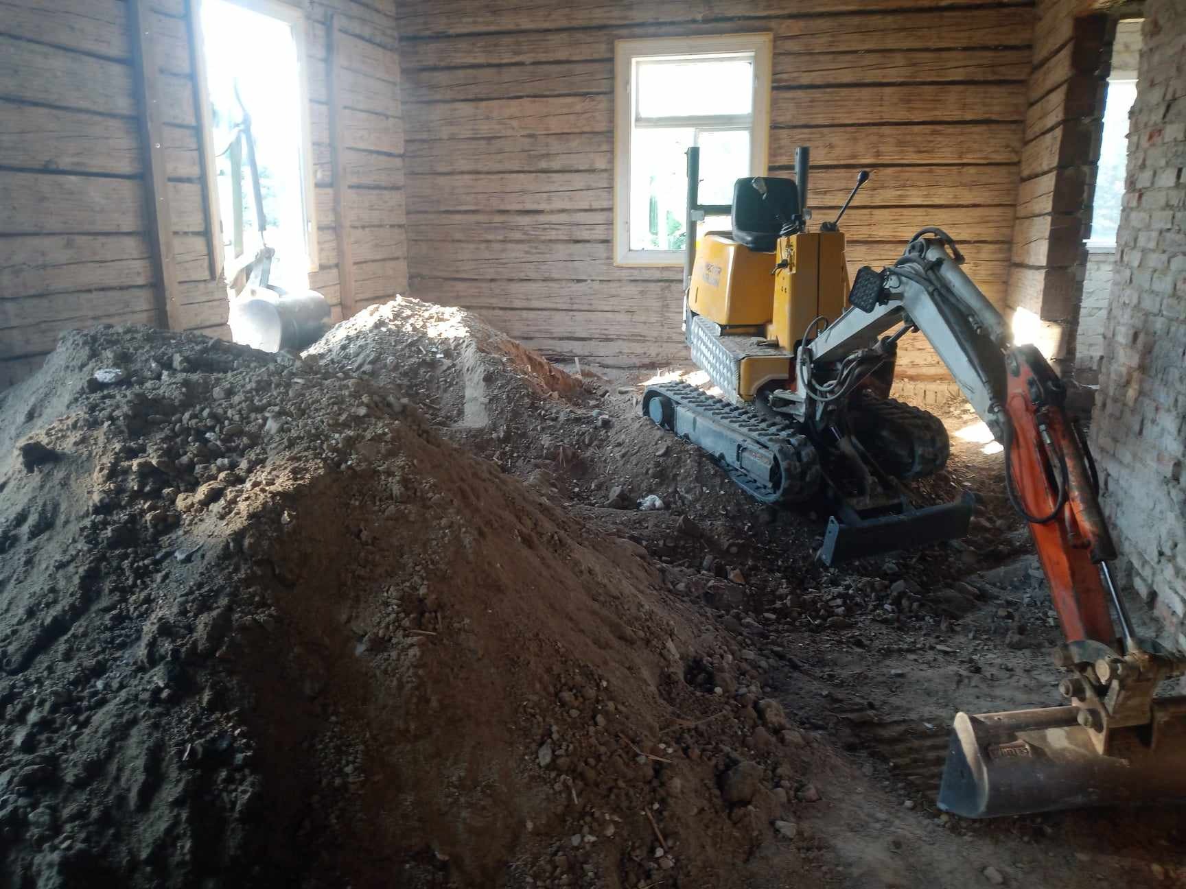 Wnętrze domu z drewnianymi ścianami w trakcie wyburzania, widoczny mały żółty i pomarańczowy koparka i sterty ziemi.