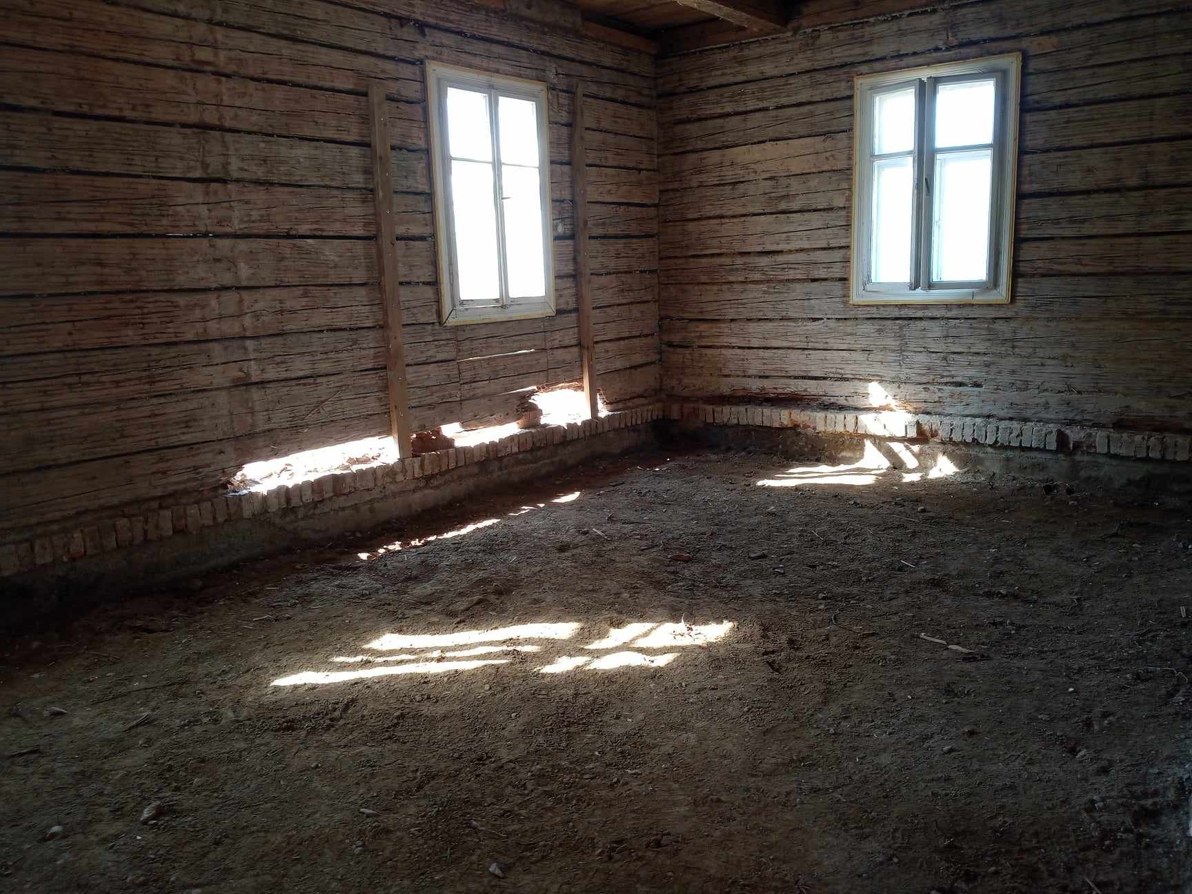 Wnętrze starego domu z drewnianymi ścianami, ceglanym fundamentem i ziemistą podłogą, oświetlone promieniami słońca wpadającymi przez okna.
