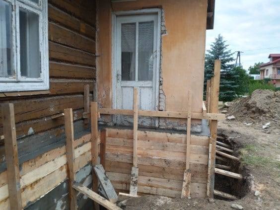 Drewniana szalunkowa konstrukcja pod fundamentem domu z bali, widoczne deski i ziemia, etap izolacji.