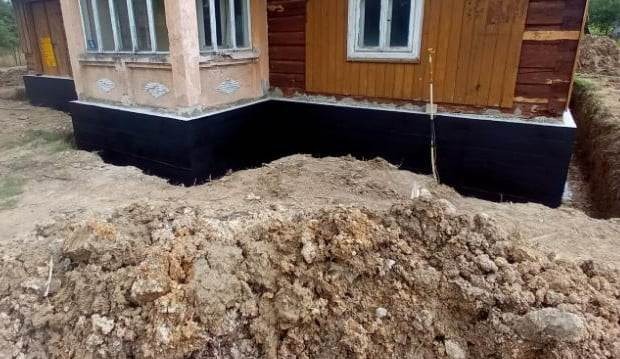 Częściowo wykopany fundament starego domu z widoczną czarną izolacją przeciwwilgociową i obróbką blacharską.
