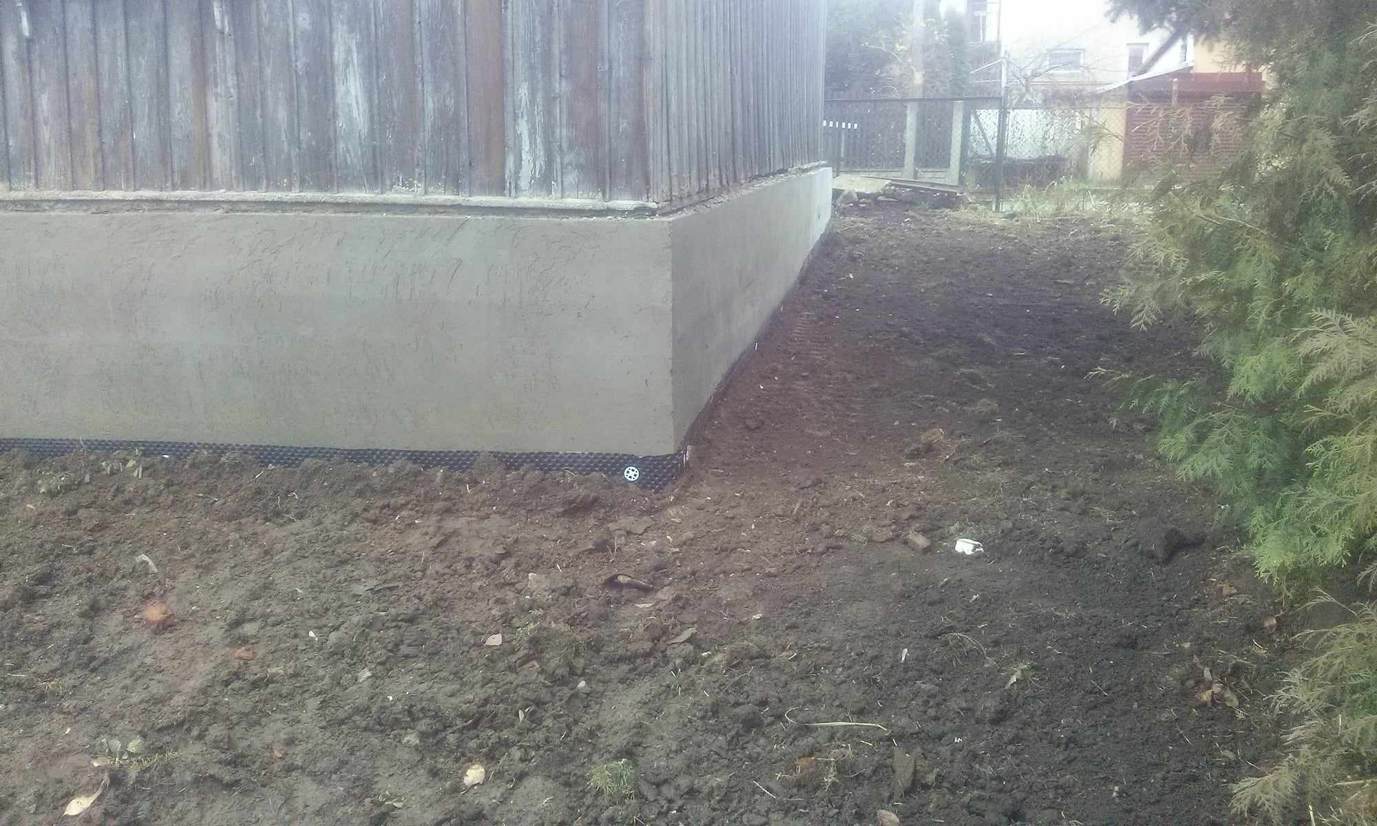 Świeżo otynkowany fragment ściany budynku z drewnianym poszyciem, widoczna siatka ochronna przy fundamencie i przygotowany teren wokół.