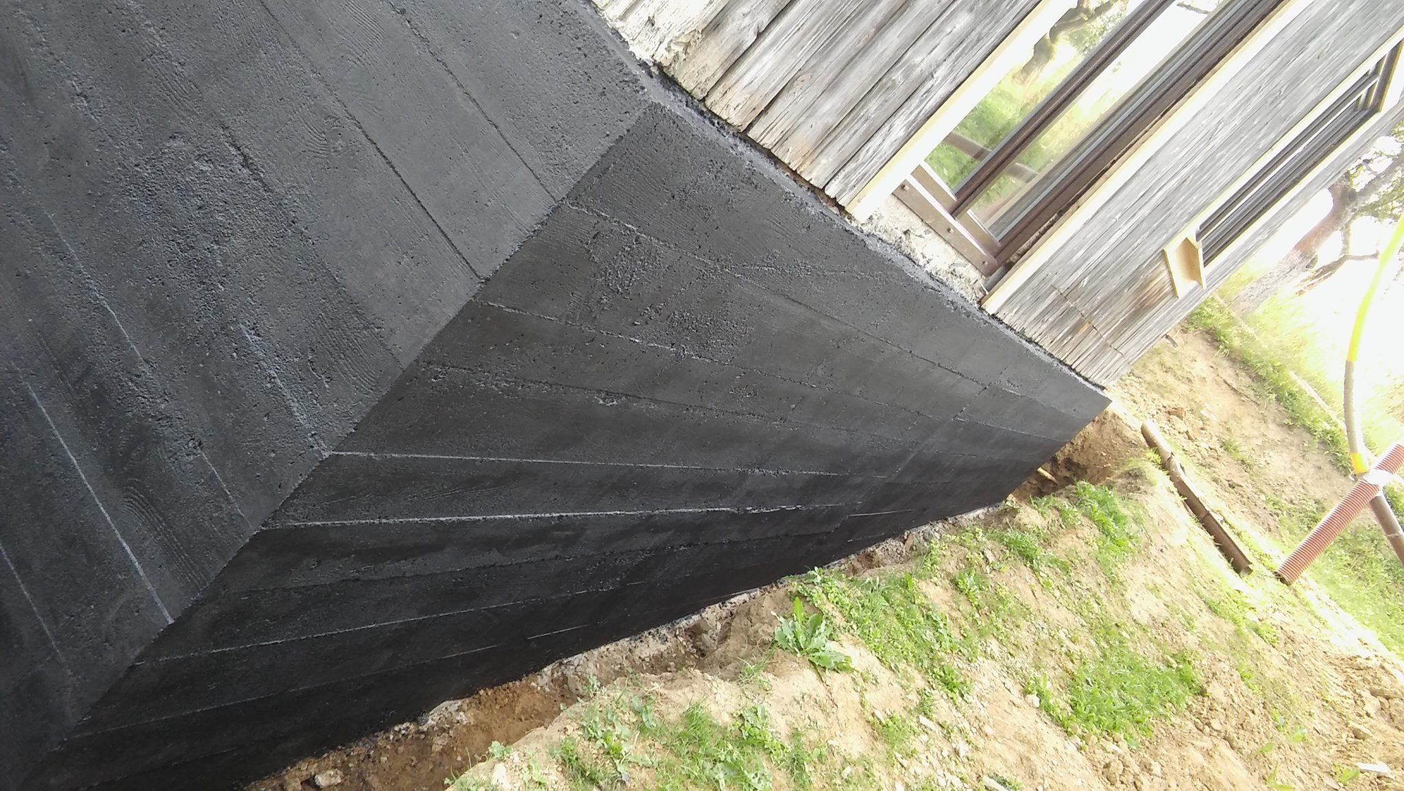 Czarna izolacja fundamentu z widoczną strukturą drewna szalunkowego, przy drewnianym budynku z oknem, obok wykop.