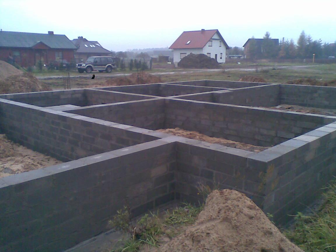 Fundamenty z bloczków betonowych na piasku, z widocznymi zarysami ścian przyszłego domu, w tle zabudowa jednorodzinna i samochód terenowy.