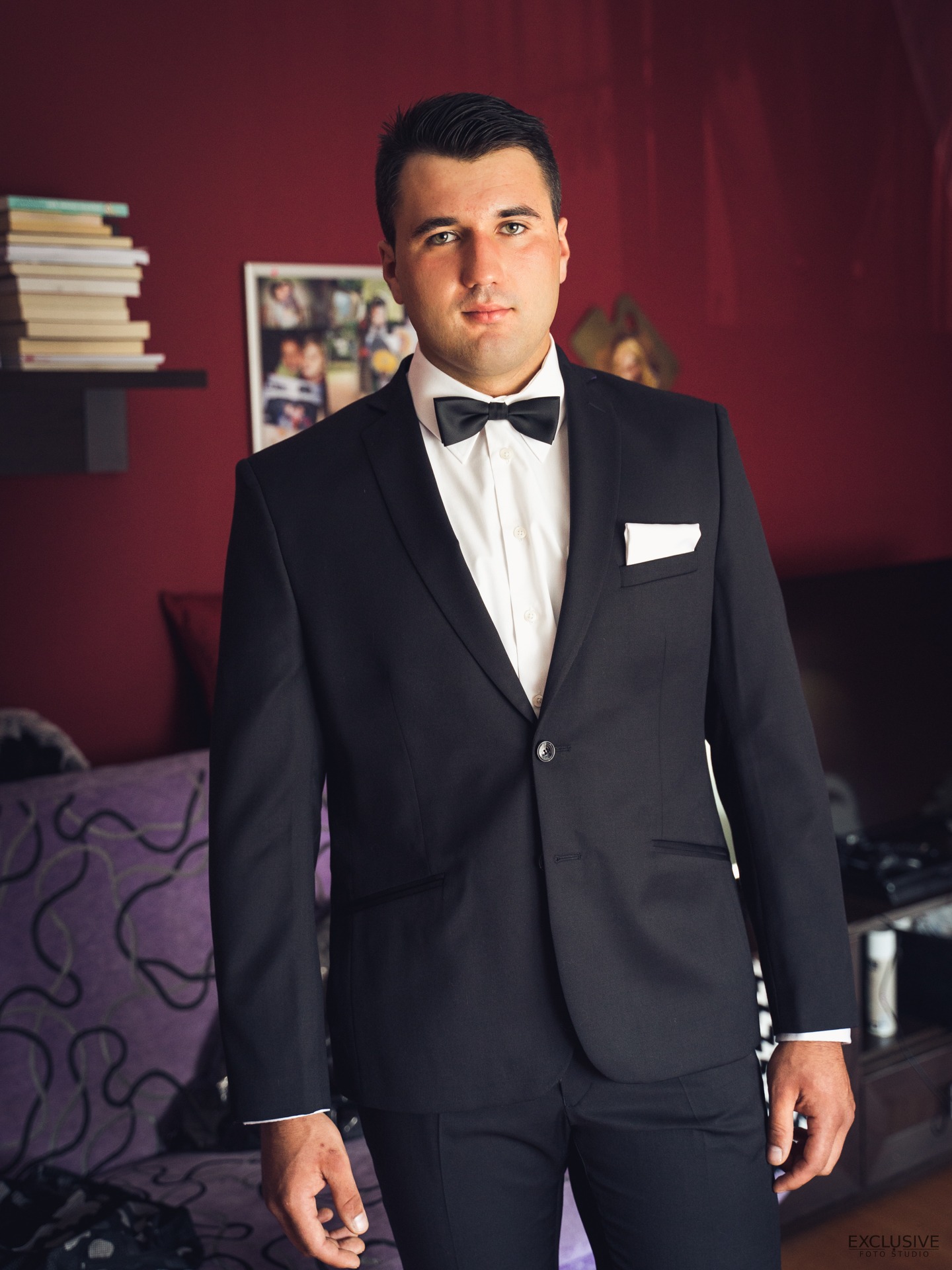 Mężczyzna w czarnym smokingu i muszce pozuje na tle bordowej ściany z półką, na której leżą książki.