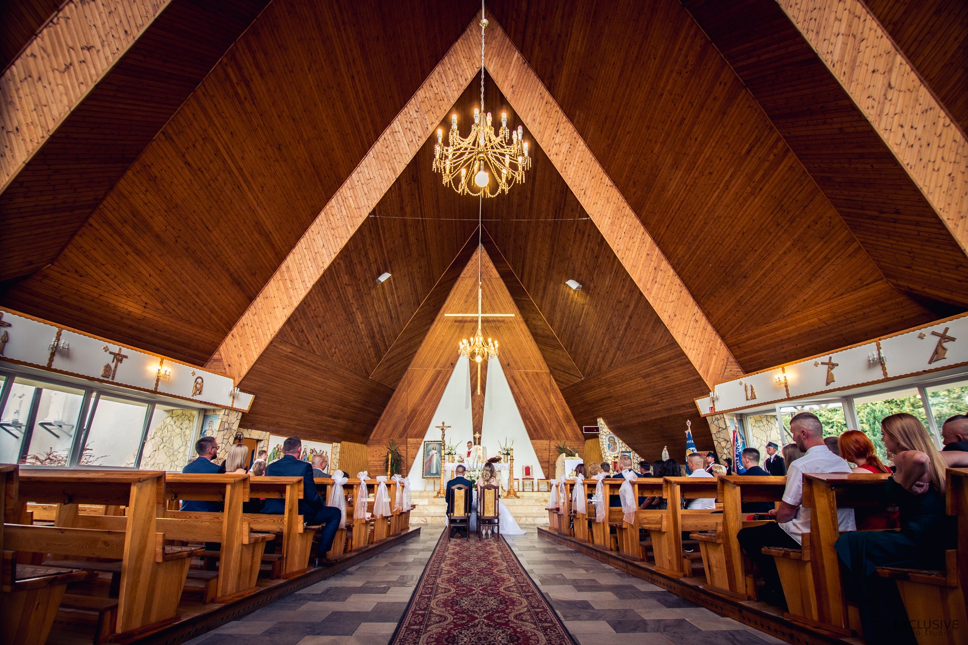 Wnętrze kościoła z drewnianym stropem w kształcie ostrosłupa, para młoda klęczy przed ołtarzem, goście siedzą w ławkach, dywan z wzorem w kolorze czerwieni prowadzi do ołtarza, dwie żyrandole wiszą...