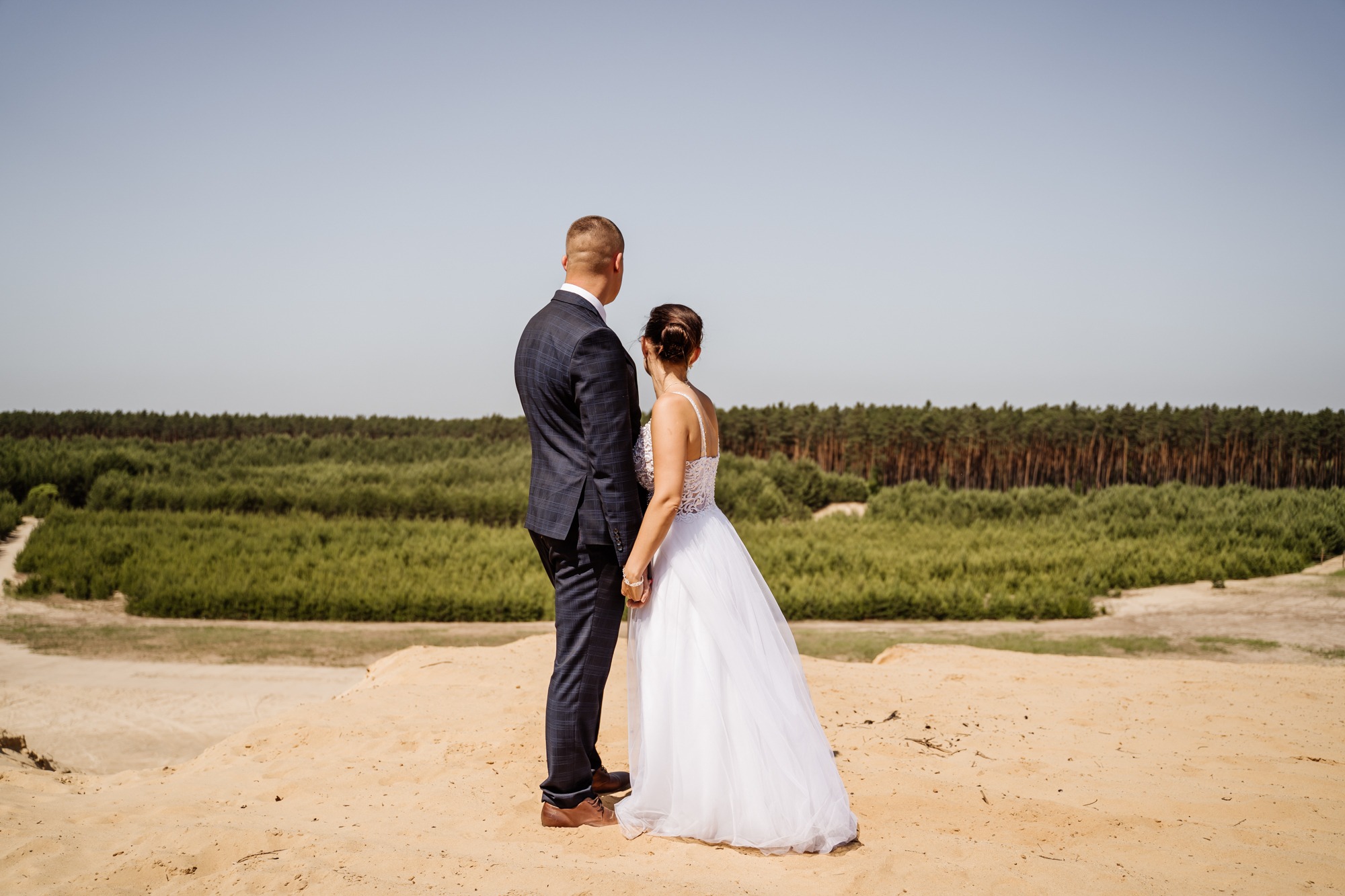 Para młoda, ubrana w granatowy garnitur i białą suknię ślubną z koronkową górą, stoi tyłem, trzymając się za ręce na piaszczystym wzgórzu z widokiem na las w tle.