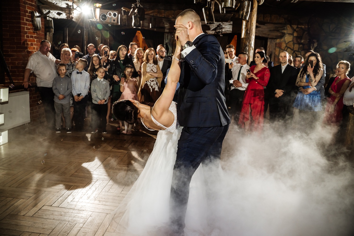 Para młoda tańczy pierwszy taniec w dymie i bańkach mydlanych, goście weselni obserwują i filmują.