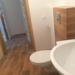 Nowoczesna łazienka po remoncie: biała umywalka i toaleta, drewnopodobne płytki na podłodze, obudowa stelaża WC w drewnie.