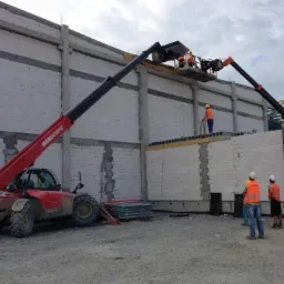 Budowa murowanego budynku w trakcie: pracownicy na podnośniku montują elementy konstrukcyjne, na dole stoją osoby w kamizelkach odblaskowych, obok czerwony podnośnik teleskopowy Maniscopic...