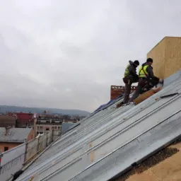 Dwóch dekarzy w kamizelkach odblaskowych montuje metalowe panele na dachu budynku w pochmurny dzień, widok na miasto w tle.