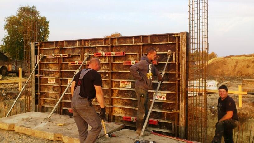 Trzech pracowników budowlanych przy szalunku fundamentów, widoczna konstrukcja zbrojeniowa, teren budowy z piaskiem w tle.