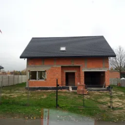 Surowy stan domu z czerwonej cegły z czarnym dachem i oknem w Radomsku, widok przez czarny metalowy płot, z widocznymi elementami konstrukcyjnymi i stosem cegieł obok wejścia.