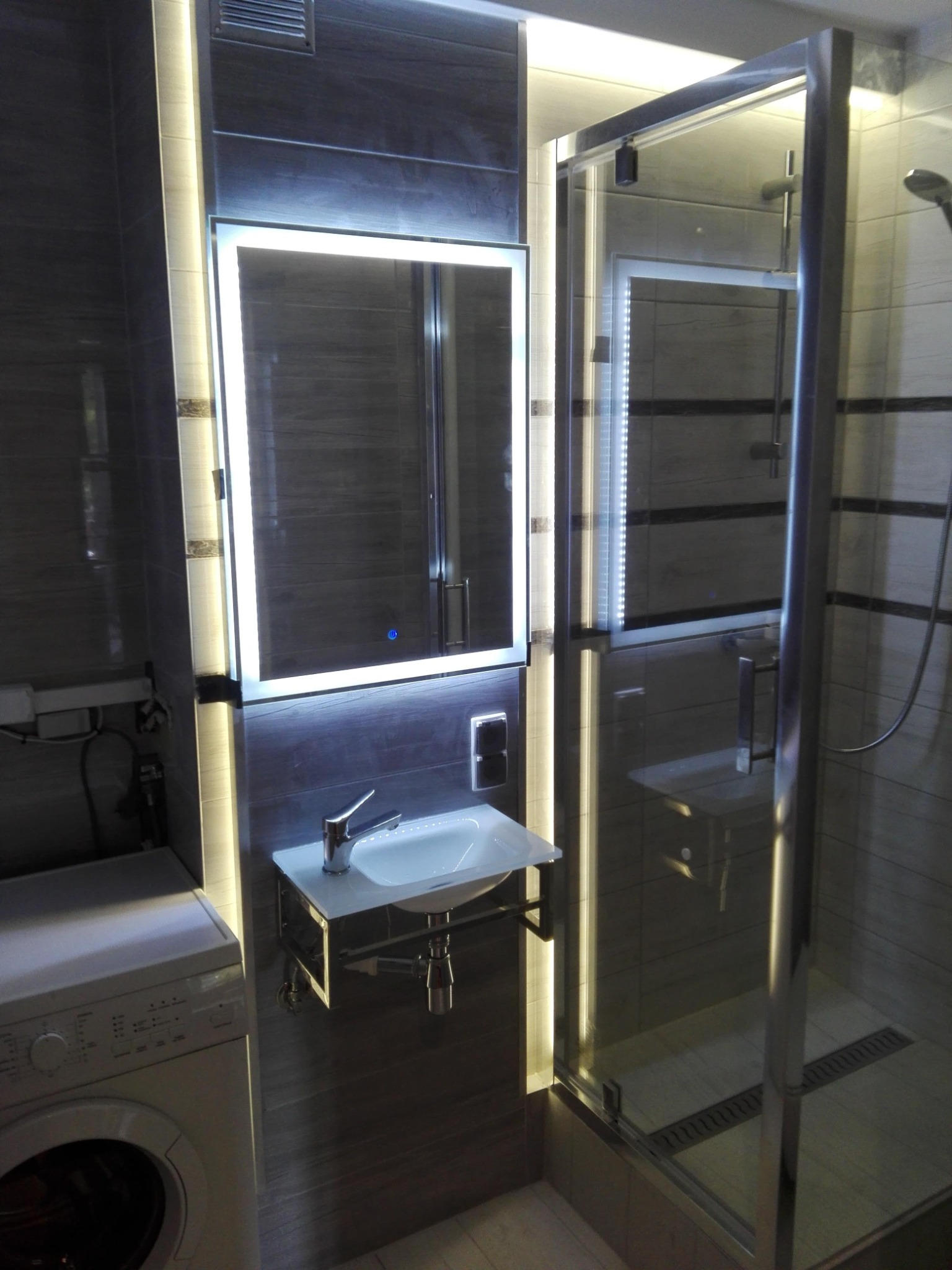 Nowoczesna łazienka z podświetlanym lustrem, szklaną kabiną prysznicową i pralką, wykończona płytkami imitującymi drewno.