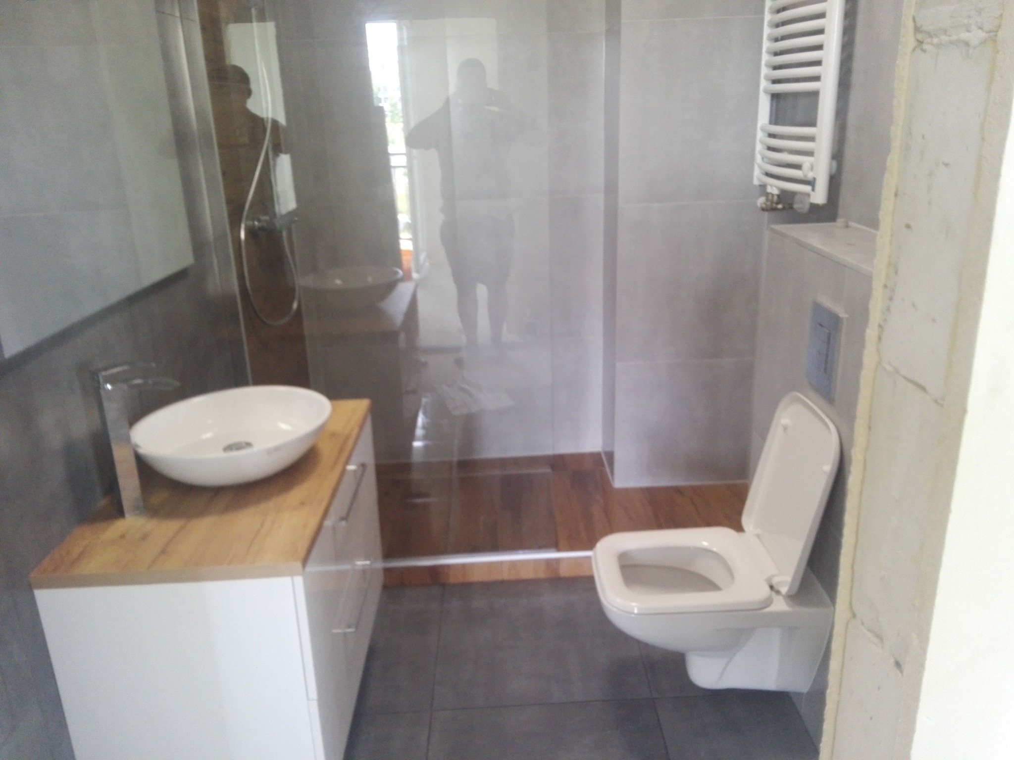 Nowoczesna łazienka z kabiną prysznicową typu walk-in, umywalką nablatową na szafce z drewnianym blatem i wiszącą toaletą, wykończona szarymi płytkami.