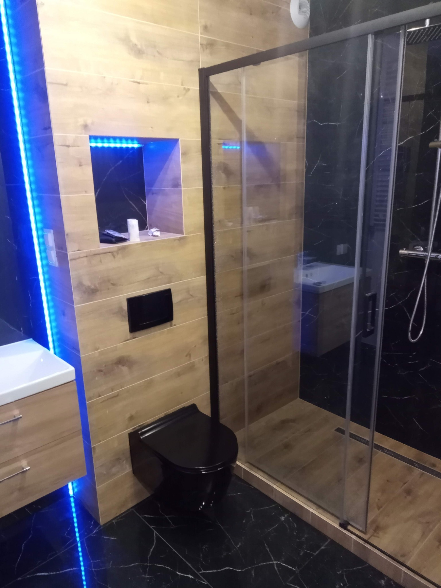 Nowoczesna łazienka z drewnopodobnymi płytkami, czarną armaturą i podświetleniem LED, kabina prysznicowa typu walk-in z czarnymi profilami.