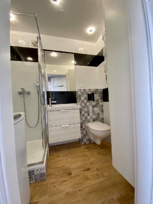 Nowoczesna łazienka z drewnopodobną podłogą, białą szafką z umywalką, kabiną prysznicową i toaletą z wzorzystą ścianą w tle.