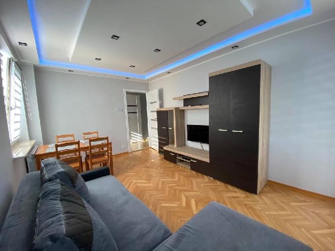 Jasne wnętrze pokoju dziennego z szarą sofą, parkietem w jodełkę, drewnianym stołem z krzesłami i zabudową meblową z telewizorem, oświetlone niebieskimi ledami na suficie.