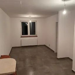 Wnętrze nowo wykończonego pokoju z brązowymi płytkami podłogowymi, białymi ścianami i grzejnikiem pod oknem. Widoczne kable sufitowe i tymczasowa żarówka.