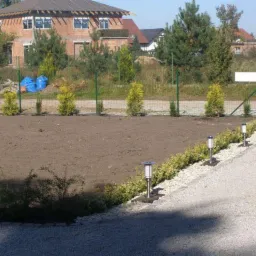 Przygotowany teren pod ogród z nasadzeniami młodych drzewek iglastych wzdłuż ogrodzenia i oświetleniem ścieżki w postaci lamp solarnych, w tle dom w trakcie budowy.