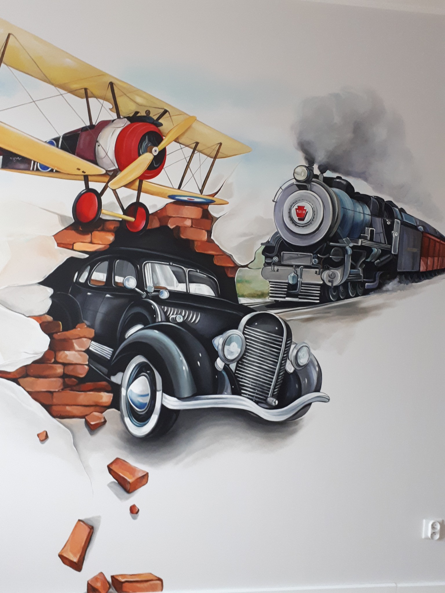 Iluzjonistyczne malowidło ścienne przedstawiające klasyczny samochód wyłaniający się z ceglanego muru, nad którym unosi się dwupłatowiec, a w tle pędzi parowóz.